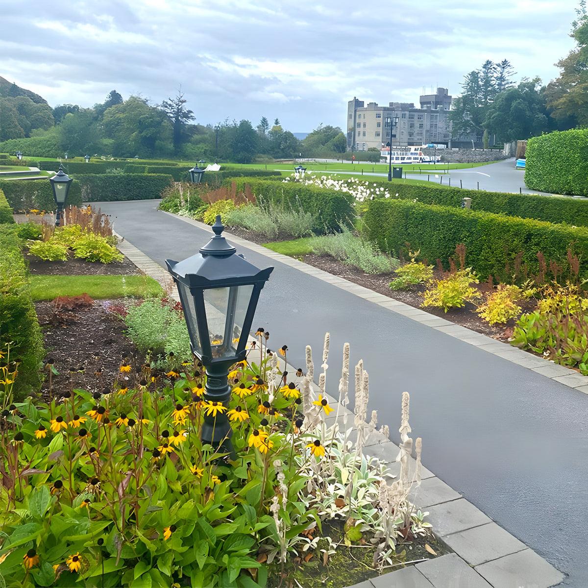 Large Victorian Cast Iron Pedestal Post Light in black, tapered four-sided glass head with stepped crown, set among garden flower beds beside a paved path.