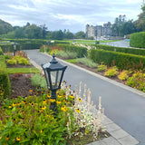 Large Victorian Cast Iron Pedestal Post Light in black, tapered four-sided glass head with stepped crown, set among garden flower beds beside a paved path.