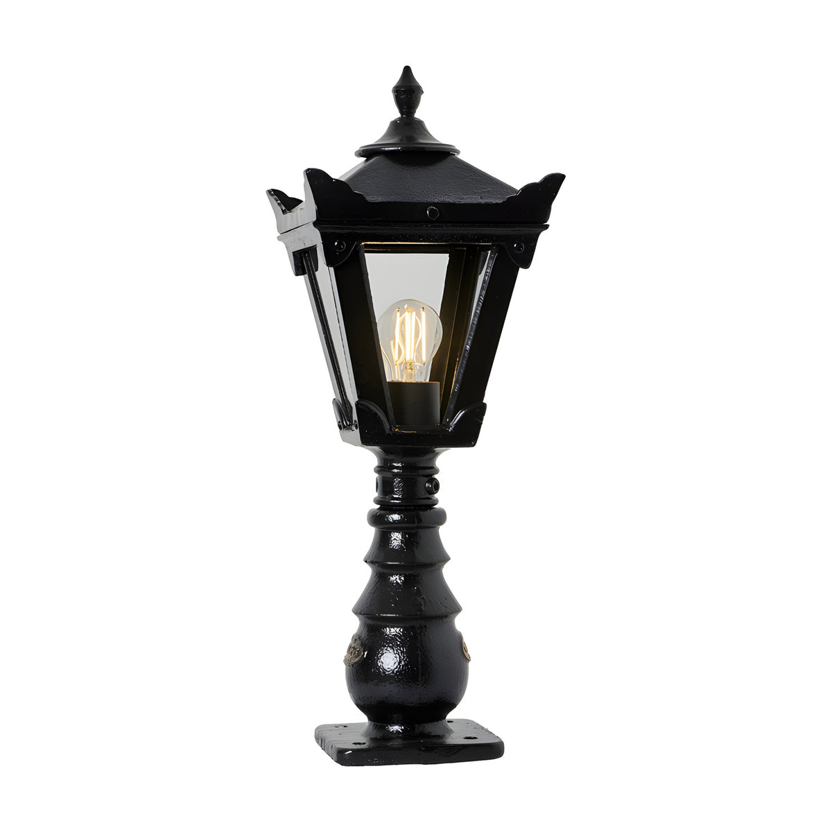 Small Victorian cast iron pedestal post light in black with a tapered four-sided glass head, stepped crown finial, and ornate base, shown lit on a plain white background.