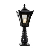 Small Victorian cast iron pedestal post light in black with a tapered four-sided glass head, stepped crown finial, and ornate base, shown lit on a plain white background.