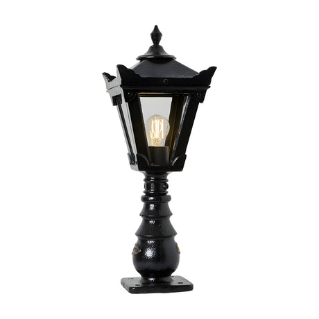 Small Victorian cast iron pedestal post light in black with a tapered four-sided glass head, stepped crown finial, and ornate base, shown lit on a plain white background.