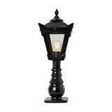 Small Victorian cast iron pedestal post light in black, tapered lantern head with stepped crown and finial, glass panels, lit bulb, on a plain white background.