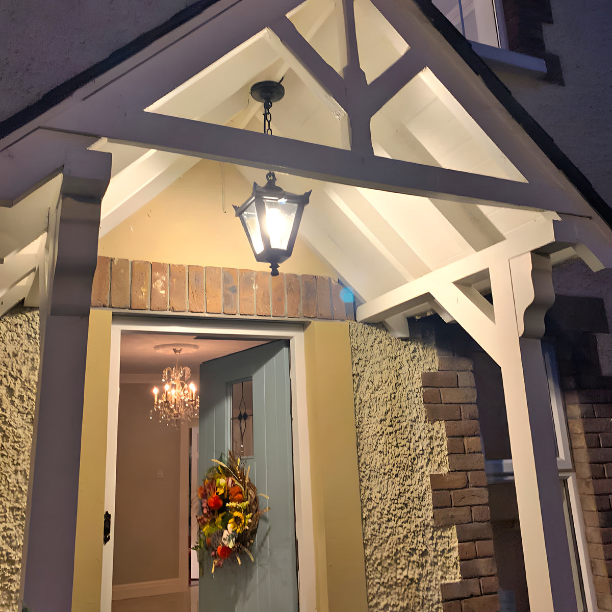 Small Victorian cast iron hanging lantern in black, four-sided glass head on a chain, illuminated beneath a porch gable at a covered entrance.