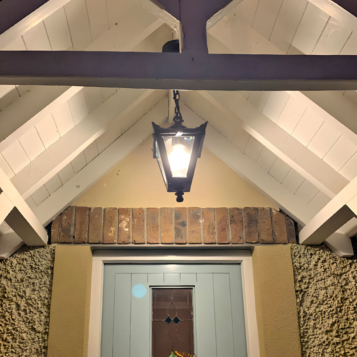 Small Victorian cast iron hanging lantern in black, tapered four-sided glass head with stepped crown, suspended on a chain beneath a white vaulted porch ceiling.