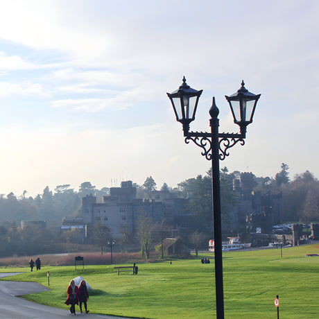 Victorian cast iron double-headed lamp post in black, 3.3m tall with two traditional lantern heads on curved brackets, shown outdoors on parkland near a castle.