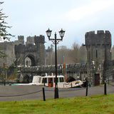 Victorian cast iron double-headed lamp post in black, 3.3m tall with two lantern heads and scrollwork bracket, installed by a castle courtyard waterside setting.