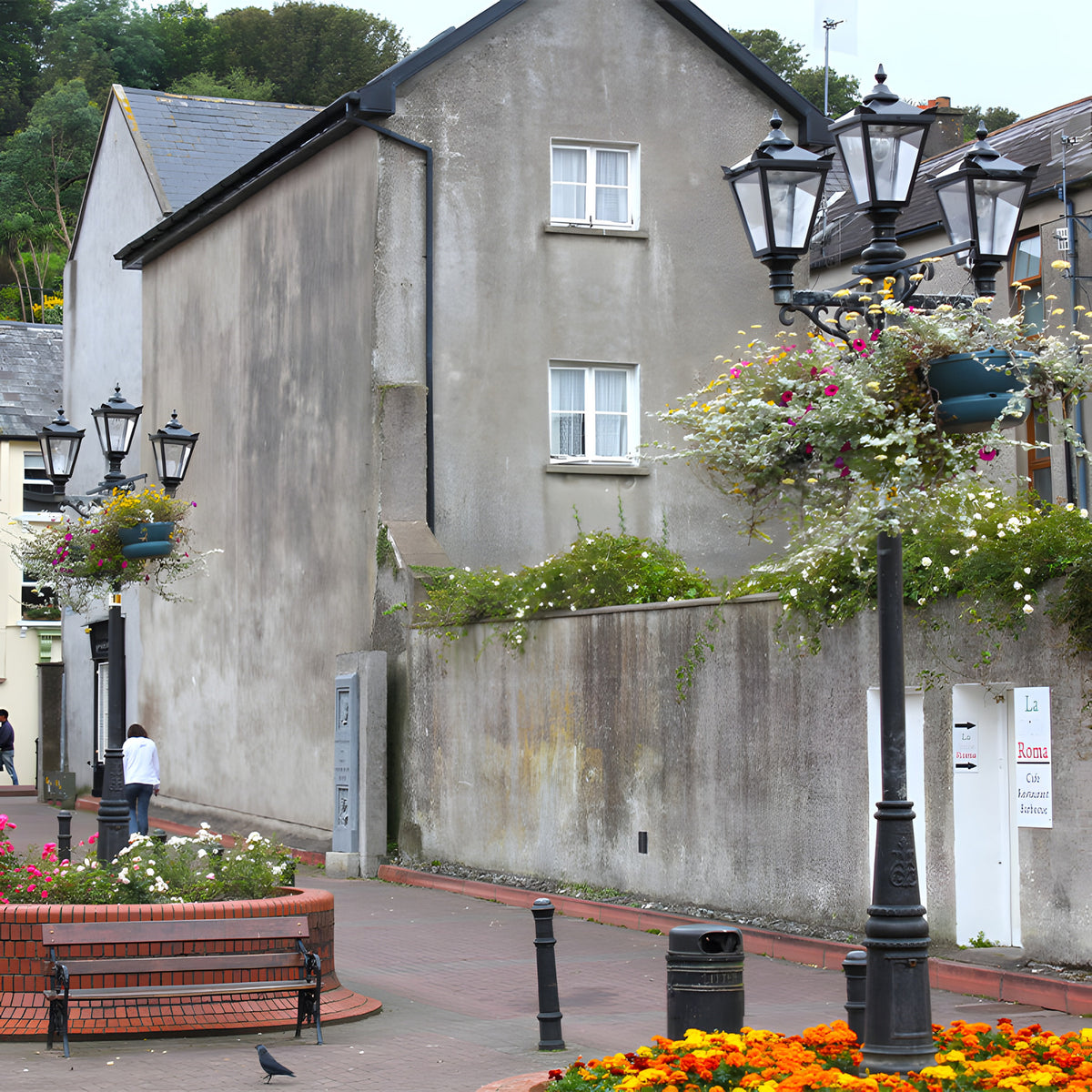 Victorian cast iron triple-headed lamp post in black, tapered lantern heads with stepped crowns, shown outdoors on a paved street lined with hanging flower baskets.