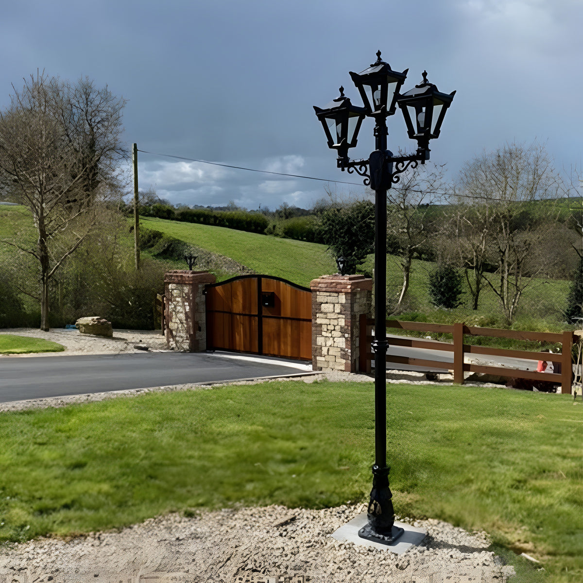 Victorian cast iron triple-headed lamp post, 2.6m black post with three lantern heads on scrolled arms, installed on a gravel base by a driveway and countryside setting.