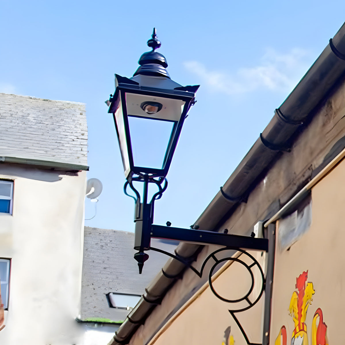 Large Victorian wall lantern in black, tapered four-sided glass head with stepped crown and finial, mounted on a black scroll bracket against a building exterior.