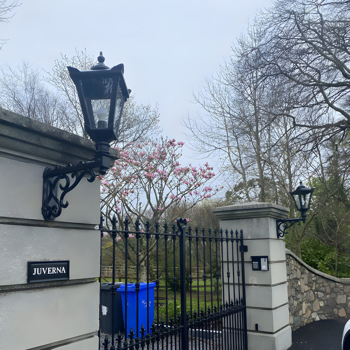 Medium Victorian wall lantern in black, tapered four-sided glass shade with cast finial, mounted on a decorative scroll bracket beside a gated entrance.