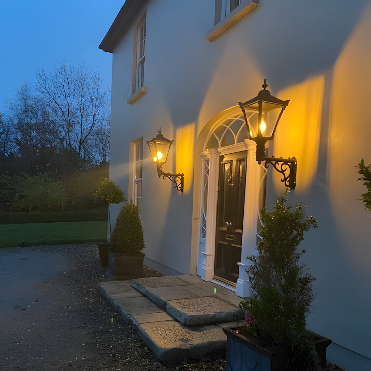 Medium Victorian wall lantern in black on a decorative cast iron scroll bracket, tapered glazed lantern illuminated beside a period front door at dusk.