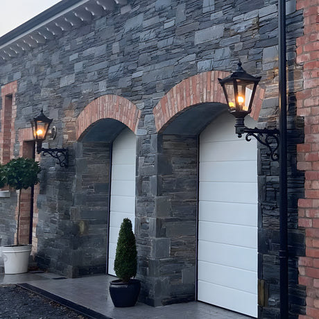 Medium Victorian wall lantern in black on a decorative cast iron scroll bracket, mounted either side of arched garage doors on a stone and brick façade, illuminated at dusk.