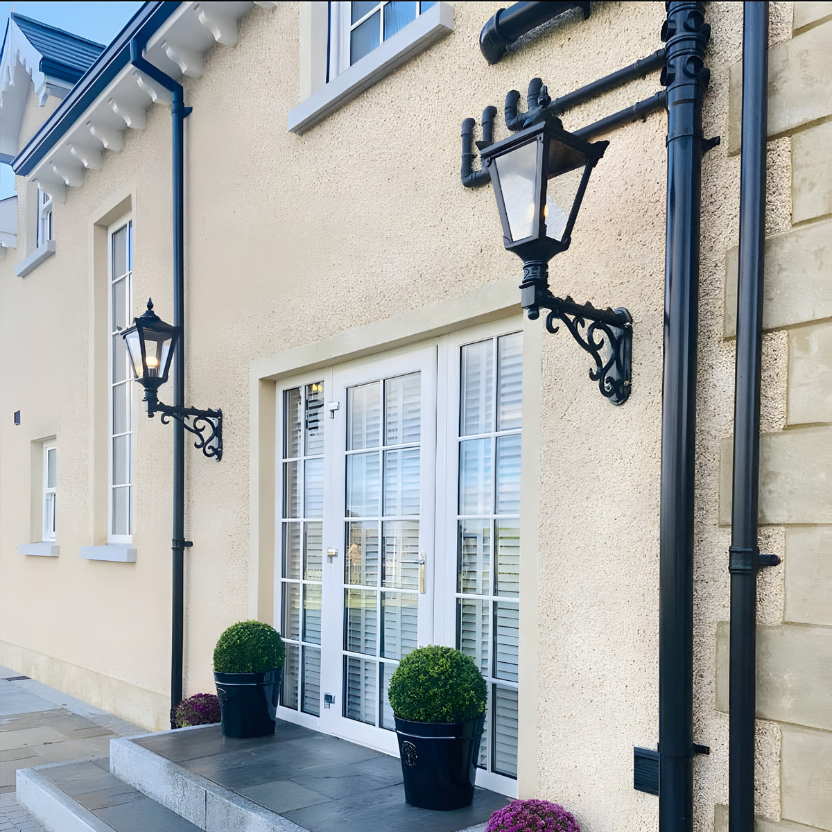 Medium Victorian wall lantern in black on a decorative cast iron scroll bracket, mounted on a house exterior beside a white-framed door with glazed panels.