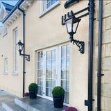 Medium Victorian wall lantern in black on a decorative cast iron scroll bracket, mounted on a house exterior beside a white-framed door with glazed panels.