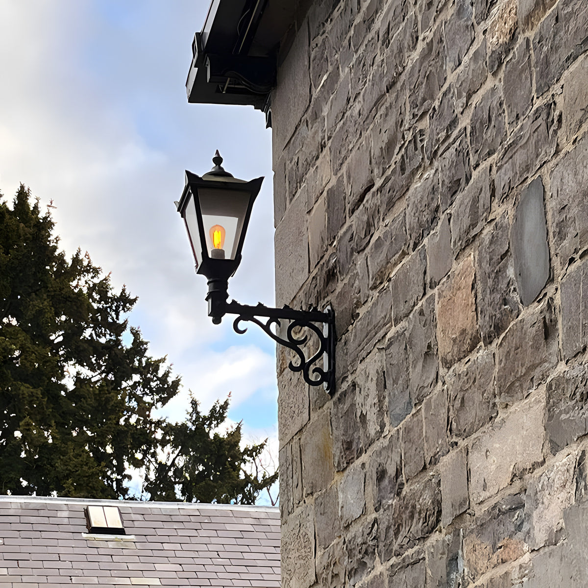 Medium Victorian Wall Lantern on a scroll bracket in black, tapered glass lantern with cast finial mounted on a stone wall exterior.