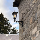 Medium Victorian Wall Lantern on a scroll bracket in black, tapered glass lantern with cast finial mounted on a stone wall exterior.