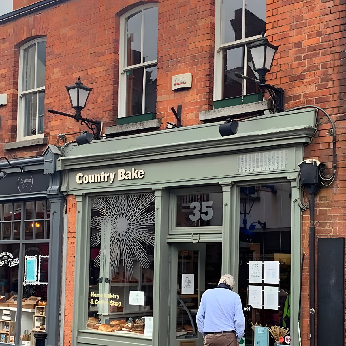 Victorian cast iron wall lantern in black, tapered four-sided glass head with stepped crown, mounted on a long black arm above a brick shopfront.