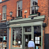 Victorian cast iron wall lantern in black, tapered four-sided glass head with stepped crown, mounted on a long black arm above a brick shopfront.