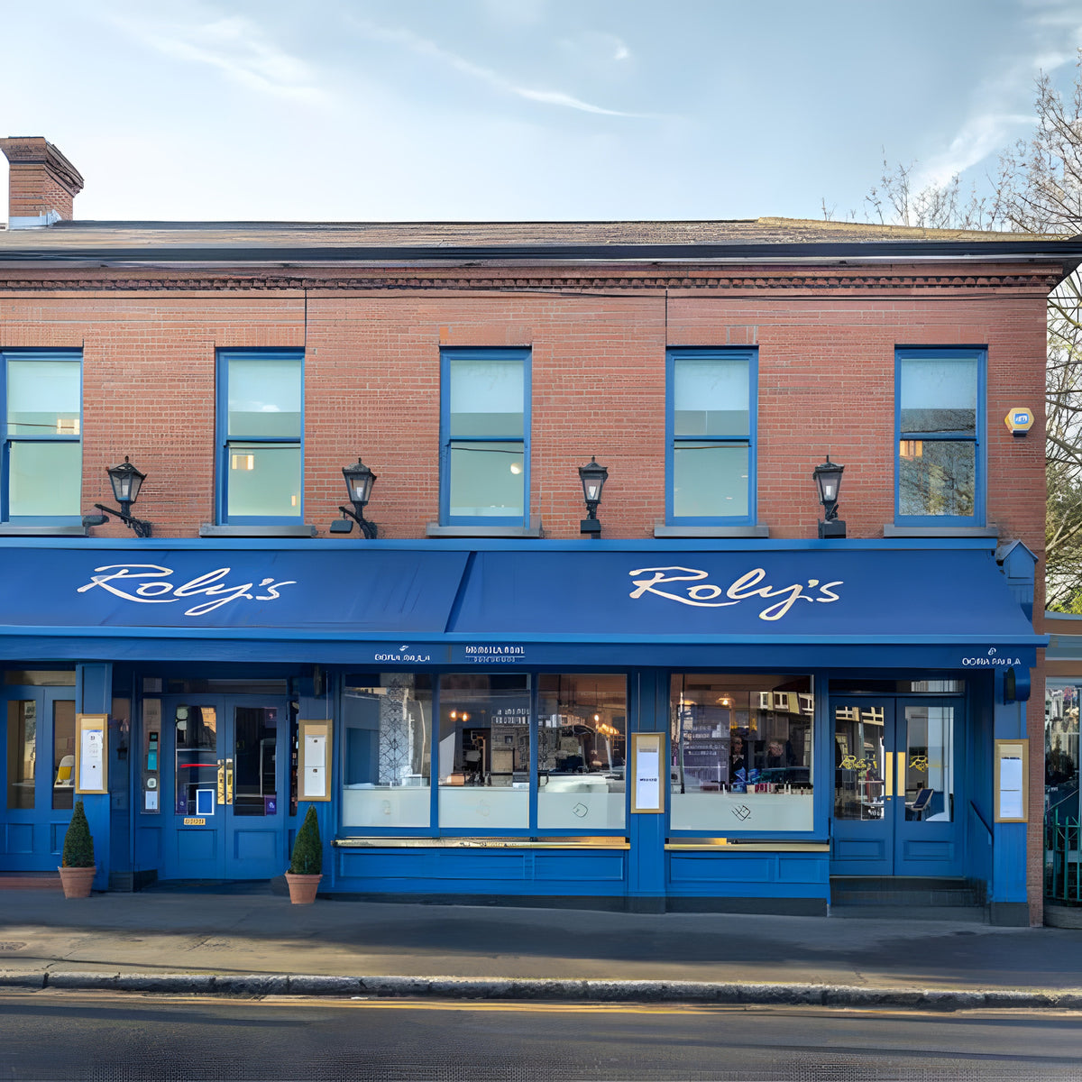 Victorian cast iron wall lantern in black, mounted on a long arm on a brick storefront, with a tapered four-sided glass head and stepped crown above blue-framed shopfront windows.