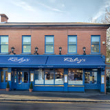 Victorian cast iron wall lantern in black, mounted on a long arm on a brick storefront, with a tapered four-sided glass head and stepped crown above blue-framed shopfront windows.