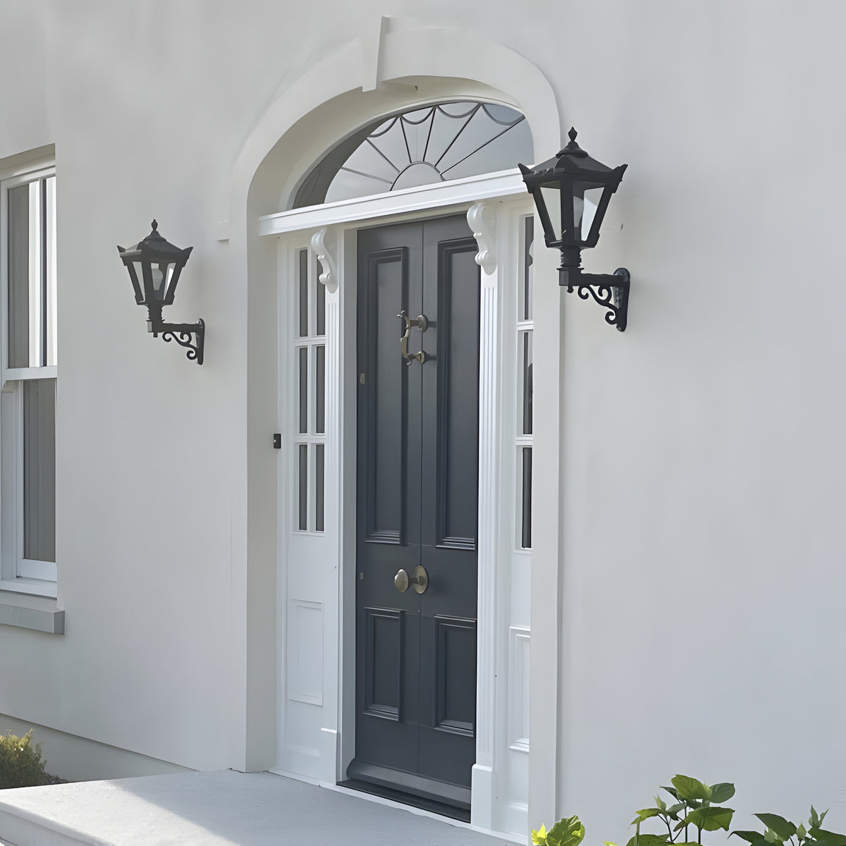 Two Small Victorian Wall Lanterns on scroll brackets in black, mounted either side of a panelled front door on a light grey house exterior.