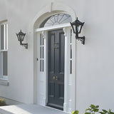 Two Small Victorian Wall Lanterns on scroll brackets in black, mounted either side of a panelled front door on a light grey house exterior.