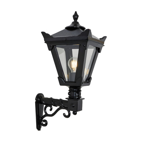 Small Victorian wall lantern in black with tapered four-sided glass head, stepped crown finial, and cast scroll bracket, shown lit on a plain white background.