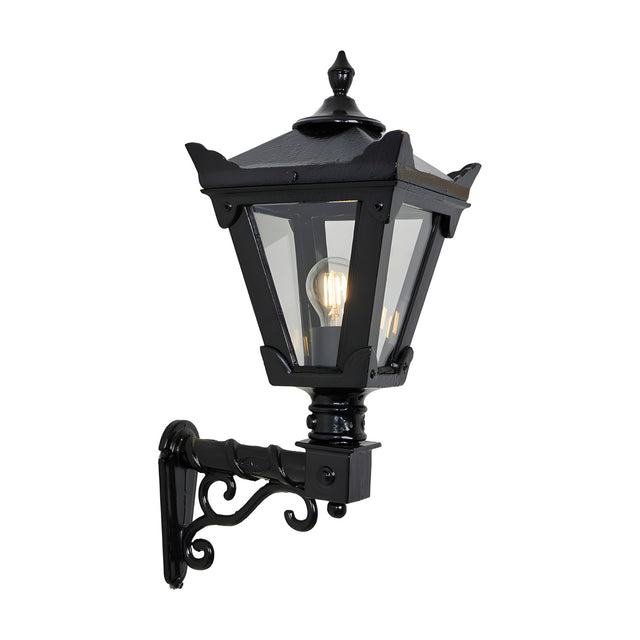 Small Victorian wall lantern in black with tapered four-sided glass head, stepped crown finial, and cast scroll bracket, shown lit on a plain white background.