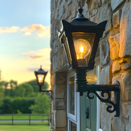 Small Victorian wall lantern in black, tapered four-sided glass head with stepped crown and finial, mounted on a scroll bracket beside a stone building entrance at dusk.
