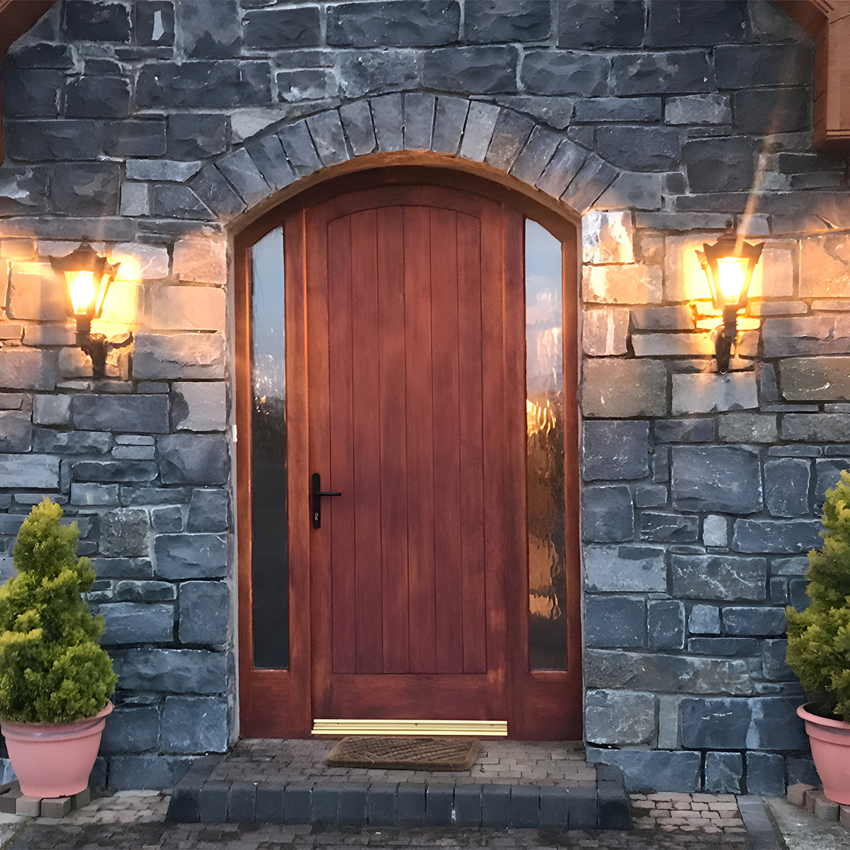 Small Victorian wall lantern in black on a scroll bracket, mounted either side of a wood front door on a stone wall, illuminated at dusk.