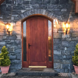 Small Victorian wall lantern in black on a scroll bracket, mounted either side of a wood front door on a stone wall, illuminated at dusk.