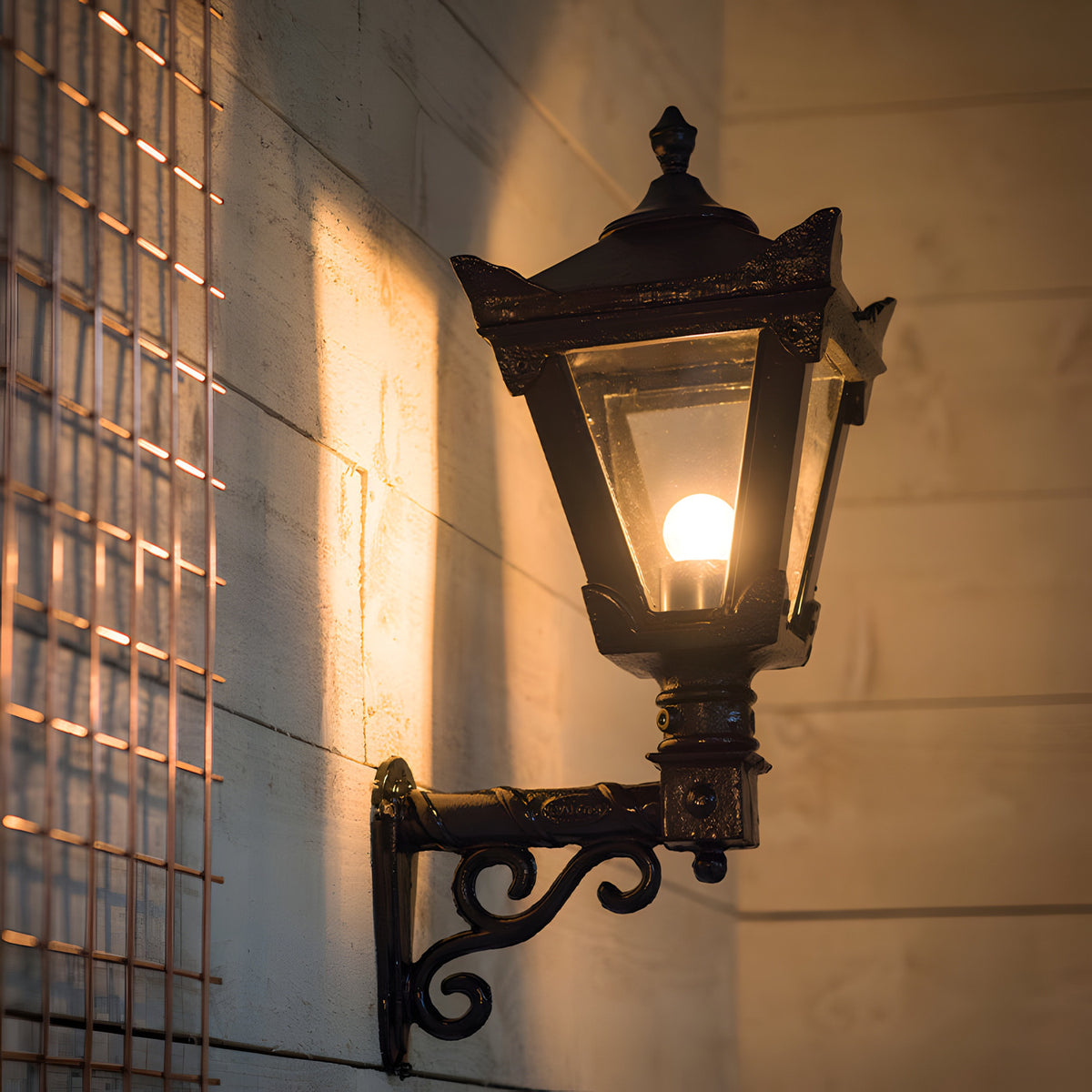 Small Victorian wall lantern in black with a tapered four-sided glass head, stepped crown, cast finial and lit E27 bulb, mounted on a scroll bracket against an exterior wall.