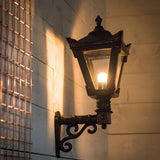 Small Victorian wall lantern in black with a tapered four-sided glass head, stepped crown, cast finial and lit E27 bulb, mounted on a scroll bracket against an exterior wall.