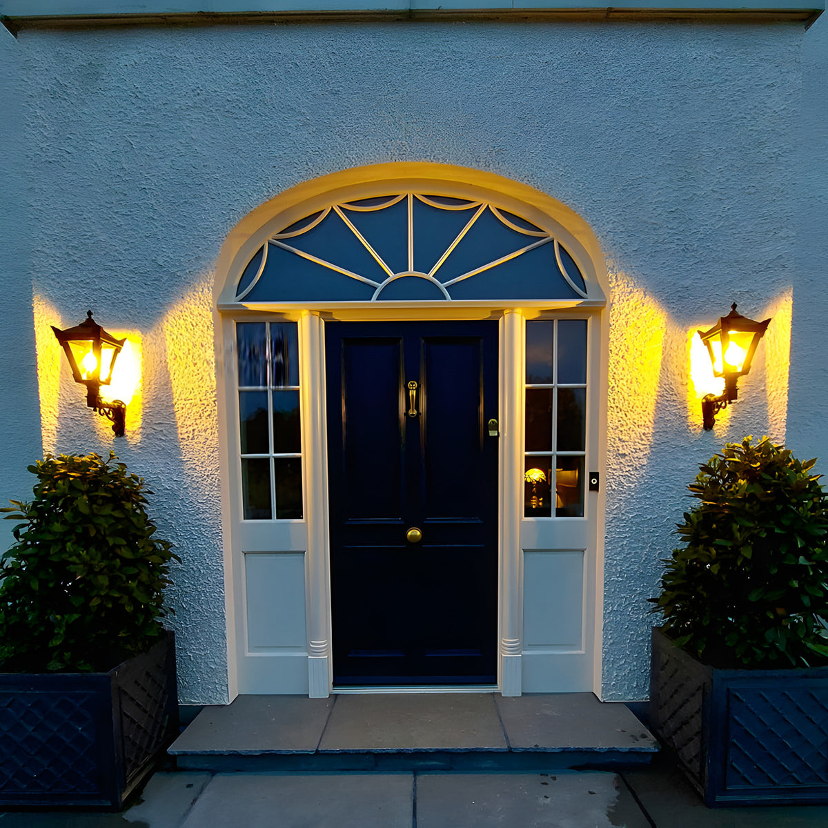 Small Victorian wall lantern in black on a scroll bracket, mounted either side of a front door with a glazed fanlight above, illuminated at dusk.