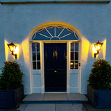 Small Victorian wall lantern in black on a scroll bracket, mounted either side of a front door with a glazed fanlight above, illuminated at dusk.