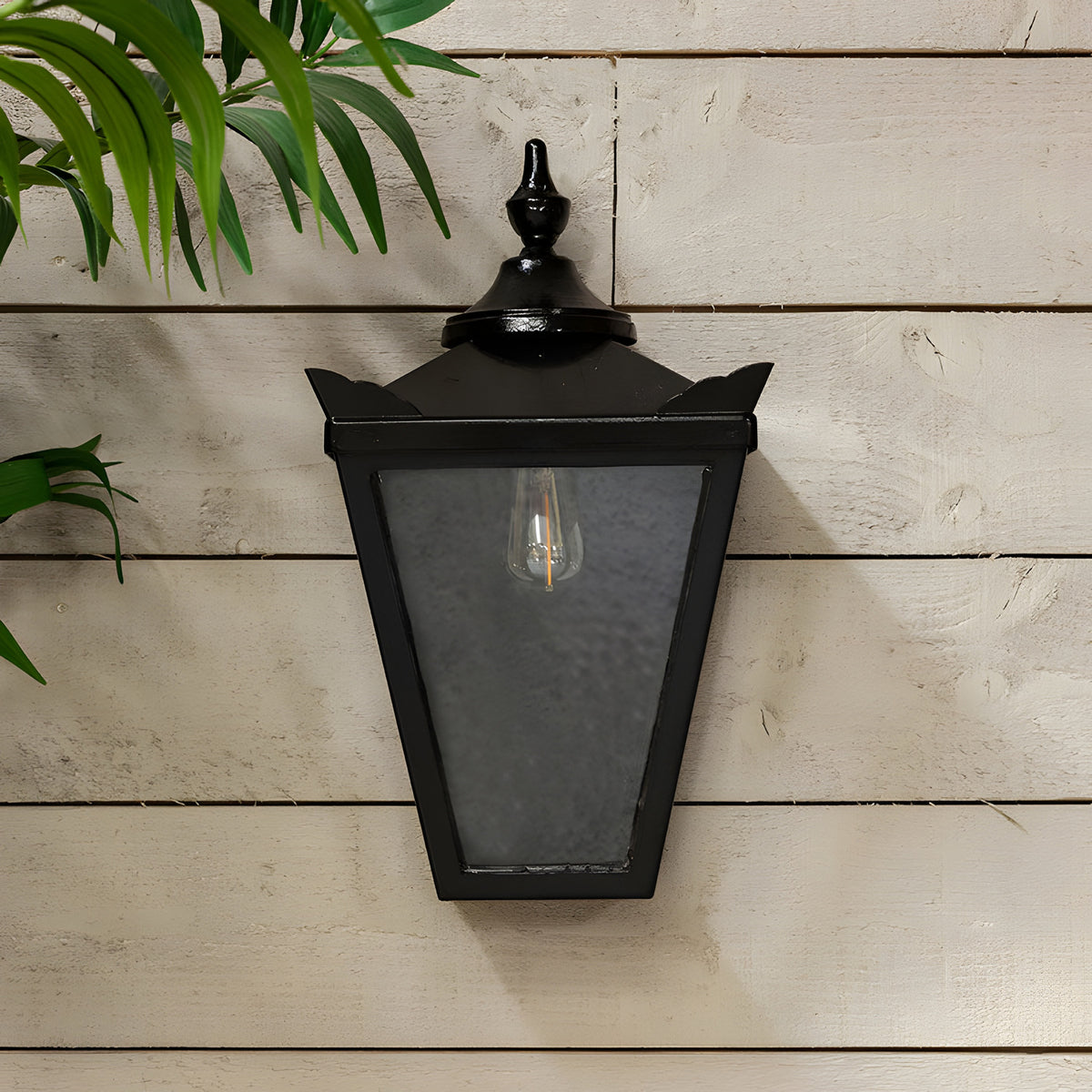 Large Victorian half lantern outdoor wall light in black cast iron with clear glass panels and an exposed E27 bulb, mounted on a white wooden panel wall.