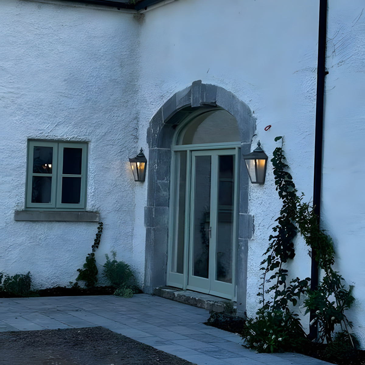 Large Victorian Cast Iron Half Lantern Wall Light in black, mounted either side of an arched front door on a white rendered house facade at dusk.