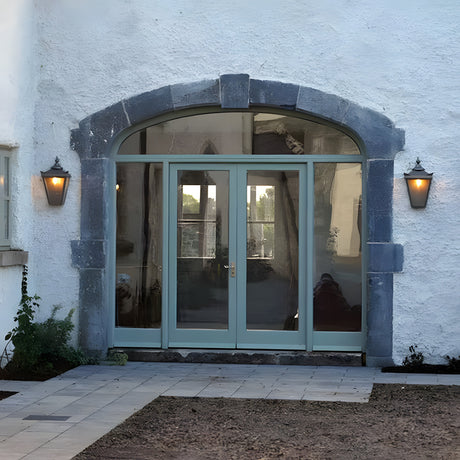 Large Victorian half lantern wall lights in black cast iron with glass panels, mounted either side of an arched glazed doorway on a white rendered exterior.