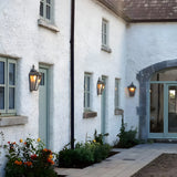 Large Victorian cast iron half lantern wall lights in black with glass panels, illuminated beside doors and windows on a white farmhouse-style exterior.