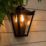 Large Victorian half lantern wall light in black, mounted on wooden cladding with clear glass panels and a lit E27 bulb visible, shown outdoors amid greenery.