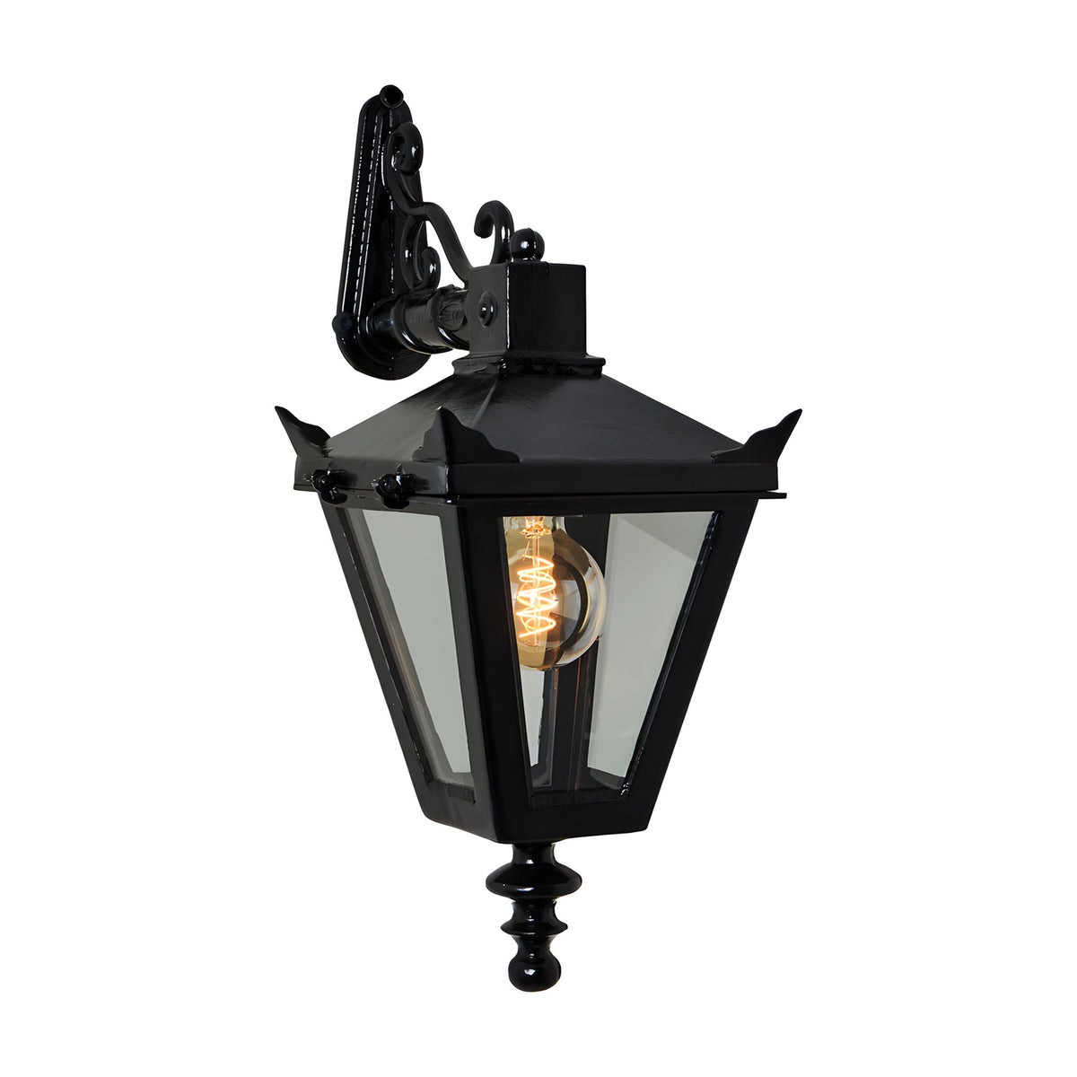 Black cast iron Victorian downturned wall lantern with tapered four-sided head, stepped crown, corner tips, clear glass panels, cast finial, and scroll bracket, shown lit on white background.