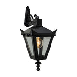 Black cast iron Victorian downturned wall lantern with tapered four-sided head, stepped crown, corner tips, clear glass panels, cast finial, and scroll bracket, shown lit on white background.