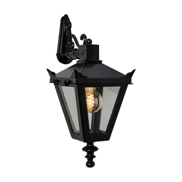 Black cast iron Victorian downturned wall lantern with tapered four-sided head, stepped crown, corner tips, clear glass panels, cast finial, and scroll bracket, shown lit on white background.