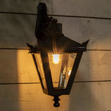 Victorian Cast Iron Downturned Wall Lantern in black, tapered four-sided glass head with ornate mounting bracket, lit and mounted on a wooden panelled wall.