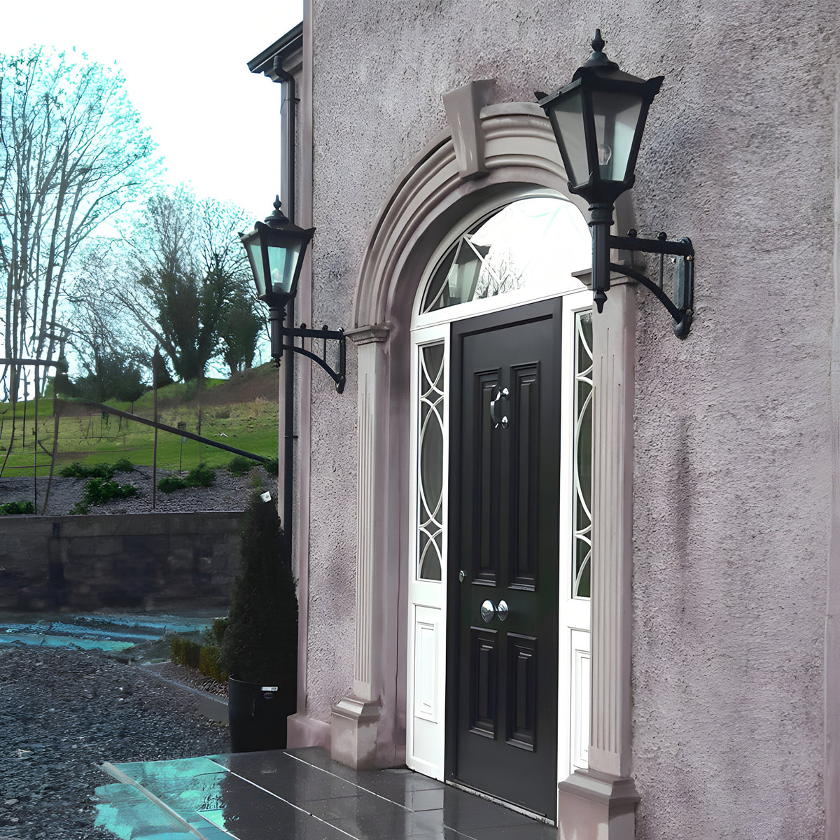 Pair of Victorian cast iron wall lanterns in black on short arms, flanking an arched front door with glass panels on a rendered house exterior.
