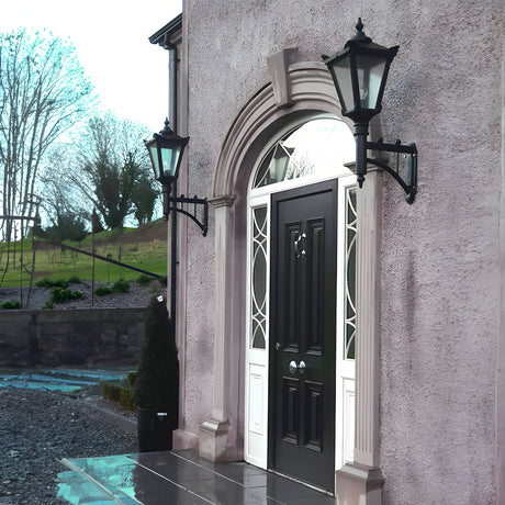 Pair of Victorian cast iron wall lanterns in black on short arms, flanking an arched front door with glass panels on a rendered house exterior.