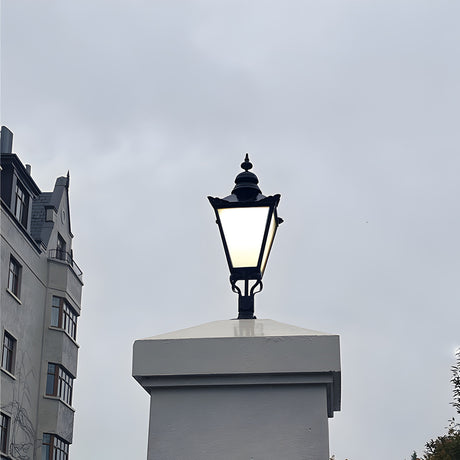Large Victorian cast iron pillar light in black, tapered four-sided lantern head with stepped crown and finial, mounted on a flat pier cap against an overcast sky.