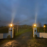 Medium Victorian cast iron pillar light in black, mounted atop flat entrance gate piers with a tapered four-sided head, stepped crown and cast finial, illuminated at dusk along a gravel driveway.