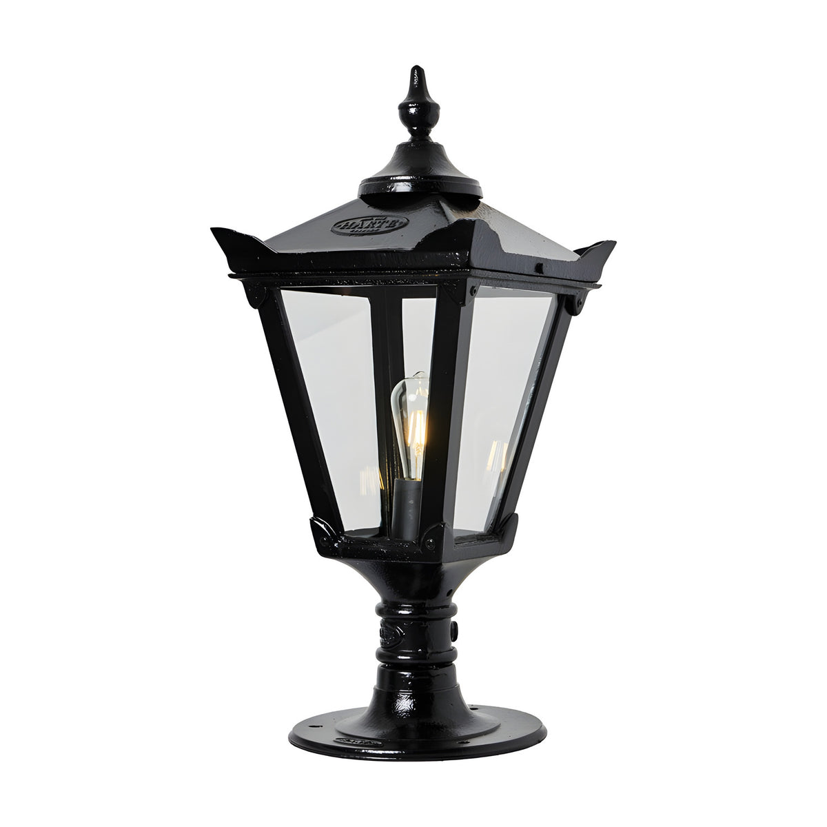 Black cast iron Victorian pillar light with tapered four-sided lantern, clear glass panels, stepped crown, corner tips, cast finial, and pier cap base, shown lit on a white background.