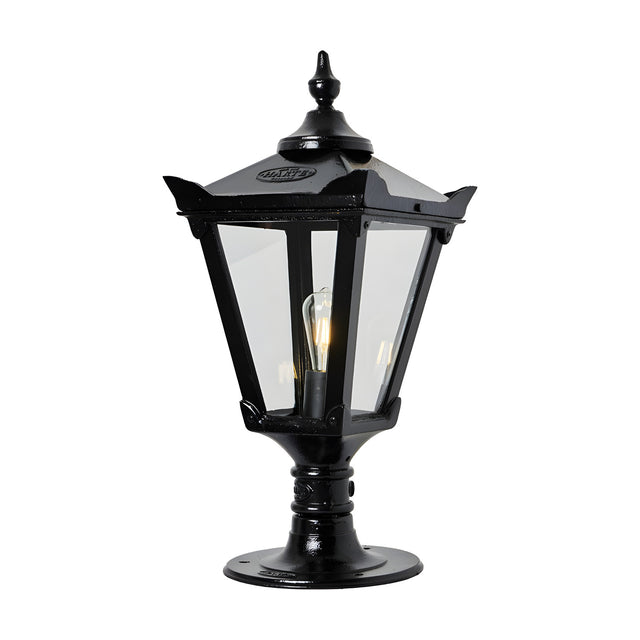 Black cast iron Victorian pillar light with tapered four-sided lantern, clear glass panels, stepped crown, corner tips, cast finial, and pier cap base, shown lit on a white background.